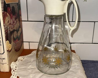May include: A clear glass pitcher featuring a white plastic lid with an integrated handle and a small pouring spout. The glass body is decorated with a repeating pattern of gold-colored starburst shapes around the center and a sunburst design on the base.