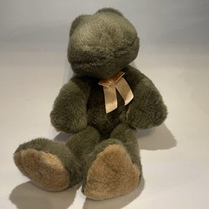 May include: A plush frog toy made of dark olive green fuzzy fabric with a soft texture. The frog has light tan fabric on the soles of its feet and a pale peach-colored satin ribbon tied in a bow around its neck. It is in a seated position.