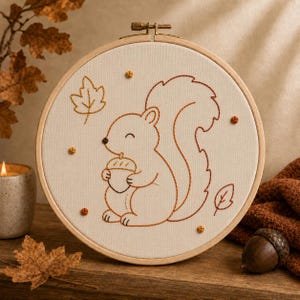 May include: A circular wooden embroidery hoop containing a cream-colored fabric panel. The fabric features a brown outline stitch design of a squirrel holding an acorn, surrounded by small embroidered leaves and scattered brown French knot accents.