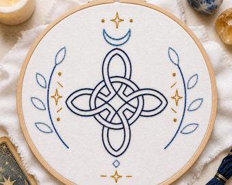 May include: A circular wooden embroidery hoop containing white fabric with a hand-stitched design. The center features a dark blue Celtic knot, flanked by two leafy branches. Above and below are blue crescent moons and small gold star accents.