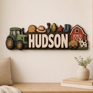 Op de afbeelding: Een decoratief wandbord met de naam HUDSON in grote witte letters. Het bord bevat een groene tractor, een hooibaal, een hoed, een schep, maïs, een rode ster, blauwe laarzen, een rode schuur, een kar met hooi en een zwart-witte koe.