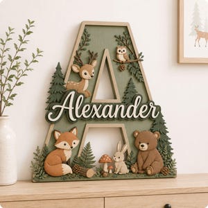 Pode incluir: Uma grande letra A decorativa de madeira em verde sálvia com o nome Alexander em letra cursiva branca. A peça é decorada com recortes de um veado, uma coruja, uma raposa, um coelho e um urso, além de pinheiros, cogumelos e pinhas.