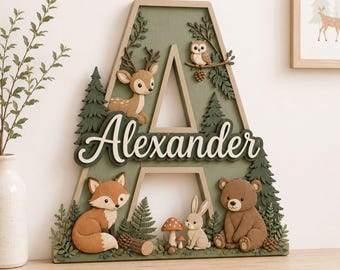Op de afbeelding: Een grote decoratieve houten letter A in saliegroen met de naam Alexander in witte schrijfletters. Het object is versierd met uitgesneden figuren van een hert, uil, vos, konijn en beer, samen met dennenbomen, paddenstoelen en dennenappels.