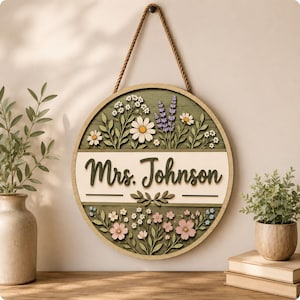 May include: A circular wooden wall sign featuring the text Mrs. Johnson in dark green script. The sign is decorated with layered, cut-out floral designs including white daisies, purple lavender, and pink flowers on a sage green background.