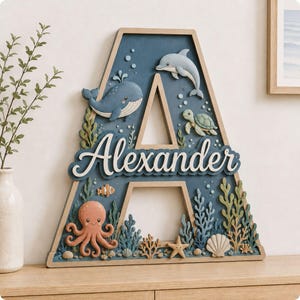 Puede incluir: Una letra A decorativa de madera con un fondo azul profundo y un tema oceánico en 3D. Incluye una ballena, un delfín, una tortuga marina, un pulpo, un pez payaso, una estrella de mar, conchas y algas. El nombre Alexander está escrito en letras blancas.
