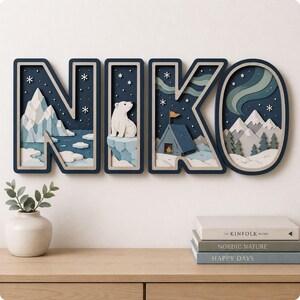 May include: A multi-layered wall art piece spelling the name NIKO in large, dark-framed letters. Each letter features a 3D winter scene with snowy mountains, a polar bear, a tent, and an aurora borealis against a dark blue starry night sky.