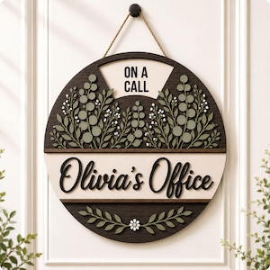 Pode incluir: Uma placa de porta circular de madeira com acabamento castanho escuro e um painel central bege claro. O texto diz ON A CALL e Olivia's Office em letras pretas. A placa é decorada com folhagem verde sálvia em camadas e pequenas flores brancas.