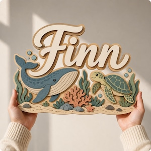 Pode incluir: Uma placa de parede de madeira em camadas com o nome Finn em letras cursivas grandes de cor creme. O design inclui uma baleia azul, uma tartaruga marinha verde, coral e algas em tons de azul, verde e pêssego sobre uma base cor de areia.