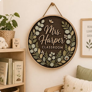 Pode incluir: Uma placa de parede circular de madeira com fundo castanho escuro, moldura de madeira clara e corda de juta para pendurar. O texto "Mrs. Harper CLASSROOM" est&aacute; em relevo, rodeado por uma coroa de folhas verdes e cor de s&aacute;lvia.