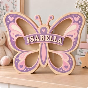 May include: A wooden butterfly-shaped piggy bank featuring a natural wood finish with purple and pink accents. The center of the butterfly displays the name ISABELLA in light-colored capital letters on a purple banner across the body.