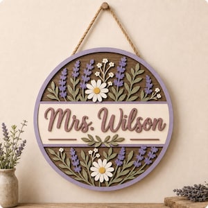 May include: A circular wooden wall sign featuring a dark brown background with a light cream horizontal center panel. The text "Mrs. Wilson" is written in a dark purple script font. The sign is decorated with purple lavender and white daisies.