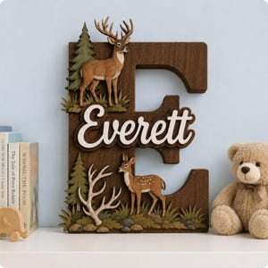 May include: A large wooden letter E featuring a layered forest scene with a brown deer, a smaller fawn, green pine trees, and white antlers. The name Everett is written in white script across the centre of the dark brown wooden letter.