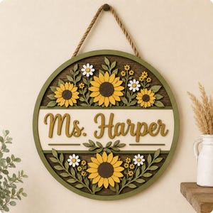 May include: A circular wooden wall sign featuring a dark brown background with layered yellow sunflowers, white daisies, and green leaves. A central cream-colored horizontal band displays the text Ms. Harper in raised, dark yellow script lettering.