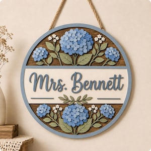 May include: A circular wooden wall sign featuring a blue frame and a rope hanger. The center displays the text Mrs. Bennett in blue script on a cream background, surrounded by layered, laser-cut blue hydrangea flowers and green leaves.