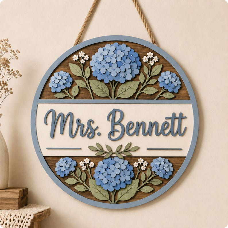 May include: A circular wooden wall sign featuring a blue frame and a rope hanger. The center displays the text Mrs. Bennett in blue script on a cream background, surrounded by layered, laser-cut blue hydrangea flowers and green leaves.