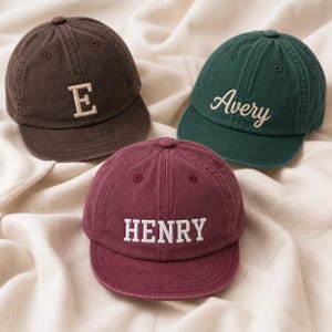 Pode incluir: Três bonés de beisebol nas cores marrom, verde-escuro e bordô. O boné marrom apresenta a letra E em bordado branco. O boné verde-escuro exibe o nome Avery em letra cursiva branca. O boné bordô mostra o nome HENRY em letras brancas de forma.