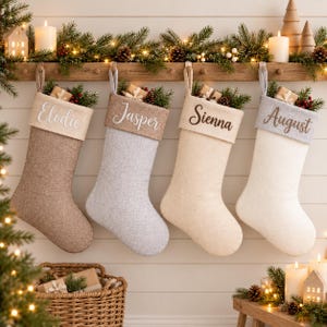 Pode incluir: Quatro meias de Natal penduradas em tons de castanho, cinzento, creme e branco. Cada meia apresenta uma bainha bordada com os nomes Elodie, Jasper, Sienna e August, preenchida com pequenas caixas de presente e ramos de pinheiro.