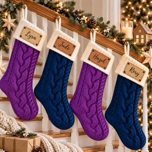 May include: Four cable-knit Christmas stockings in alternating purple and navy blue colors. Each stocking features a cream-colored cuff with a rectangular leather-like tag engraved with the names Lyra, Jude, Hazel, and Rory respectively.