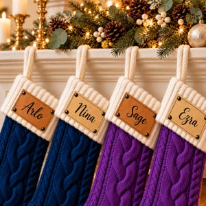 May include: Four cable-knit Christmas stockings with cream-colored cuffs and hanging loops. Two stockings are dark blue and two are purple. Each cuff features a rectangular tan leather tag secured with four black rivets, personalized with the names Arlo, Nina, Sage, and Ezra in black script.