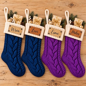 Op de afbeelding: Vier kerstsokken van kabelgebreid materiaal met crèmekleurige boorden, elk voorzien van een naamlabel in lederlook met de namen Wren, Levi, Esme of Miles. Elke sok is versierd met een klein cadeautje, dennentakjes en gouden ornamenten.