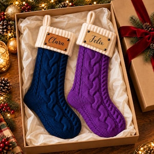 May include: Two cable-knit Christmas stockings in a gift box. One stocking is dark blue and the other is purple. Both feature a cream-colored ribbed cuff with a rectangular leather-like tag attached by four rivets. The tags are engraved with the names Clara and Felix.