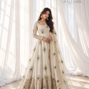 Könnte beinhalten: Ein bodenlanges, cremefarbenes Kleid mit langen Ärmeln, quadratischem Ausschnitt und einem aufwendig bestickten goldenen Saum. Das Kleid ist mit verstreuten Pfauenfeder-Motiven in Grün- und Blautönen verziert und wird mit einem passenden transparenten Dupatta kombiniert.