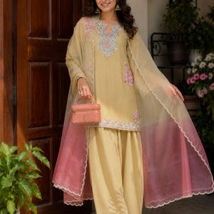 May include: A light yellow tunic and matching wide-leg trousers set featuring intricate silver embroidery around the neckline and hem. A sheer, light yellow and pink ombre dupatta with a scalloped edge is draped over the shoulders.
