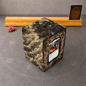 Op de afbeelding: Een vierkante deckbox met een donkere, gedetailleerde illustratie van een schedel en stormachtige wolken. Een ruilkaart wordt getoond in een transparant venster. Een rood-witte veelzijdige dobbelsteen ligt aan de linkerkant van de box.