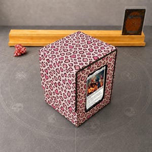 May include: A square card storage box featuring a pink and black leopard print pattern with heart-shaped spots. The front panel includes a clear display window holding a trading card with a colorful illustration and printed text.