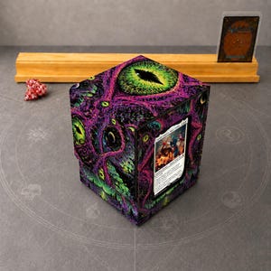 May include: A square trading card deck box featuring a vibrant purple and green cosmic eye pattern with tentacles. A single trading card is displayed in a clear front window. A red and white multi-sided die sits nearby.
