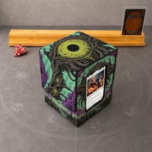 May include: A square deck box featuring a large, detailed green eye graphic on top and dark, twisted tree-like illustrations on the sides with purple accents. A single trading card is displayed in a clear window on the front side.