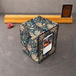 May include: A square deck box featuring a dark blue background with a repeating pattern of gold and teal coral, seaweed, and seashells. The front panel displays a single trading card in a clear window, showcasing a colorful character illustration.