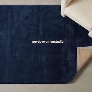 May include: A rectangular navy blue area rug with a soft, plush texture. The rug features the text wovebywomenstudio printed in white in the centre. One corner of the rug is folded back to reveal a light tan, woven fabric backing material.