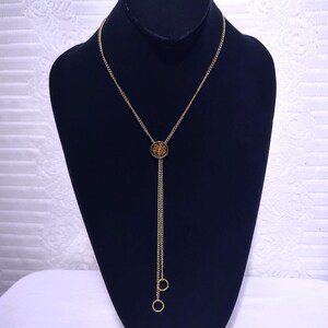 May include: A gold-toned lariat necklace featuring a thin chain that passes through a circular pendant with a black center and a gold emblem. The chain extends downward, ending in two small, open gold-toned rings at different lengths.