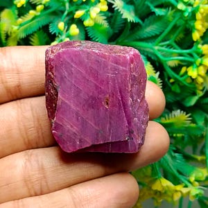 May include: A raw, uncut ruby gemstone with a deep magenta-red colour and a blocky, irregular shape. The surface of the stone displays natural striations, visible crystalline textures, and a matte finish with subtle variations in colour intensity.