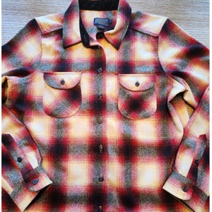 May include: A long-sleeved button-down shirt featuring a plaid pattern in shades of red, black, and cream. The shirt includes a pointed collar with black trim, two chest pockets with buttoned flaps, and buttoned cuffs. A Pendleton label is visible.