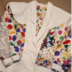 May include: A white leather jacket featuring a multi-colored patchwork design on the sleeves and front panels. The pattern includes vibrant red, yellow, green, and blue circular shapes, floral motifs, and black and white polka dots.