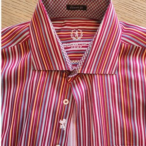May include: A long-sleeve button-down shirt featuring vertical stripes in shades of red, white, yellow, and blue. The collar interior has a light blue and white gingham pattern. The text BUGATCHI UOMO and a lion emblem are printed on the inner neck.