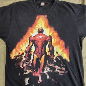 May include: A black short-sleeved t-shirt featuring a graphic print of Iron Man in a red and gold suit standing amidst a large explosion of orange and yellow flames. The Marvel logo is printed on the interior neck label of the garment.