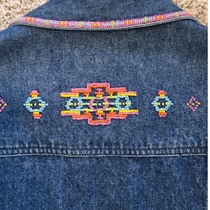 May include: A dark blue denim jacket featuring a horizontal row of intricate beadwork across the back. The embroidery consists of a central geometric pattern flanked by smaller diamond shapes in vibrant pink, yellow, and light blue beads.