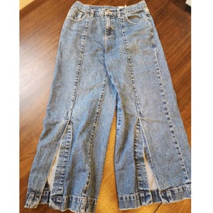 Può includere: Jeans in denim blu a lavaggio medio con vita alta, chiusura con bottone e cerniera, e cuciture verticali lungo la parte anteriore di ogni gamba. L'orlo di ogni gamba presenta un'evidente apertura verticale a fessura al centro.