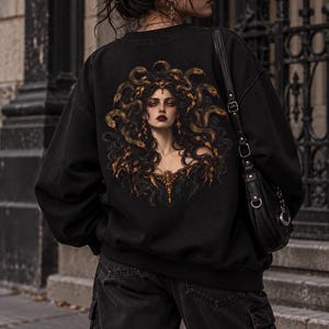 Peut inclure: Un sweat-shirt noir à manches longues avec une grande impression graphique détaillée dans le dos. L'image représente une silhouette aux cheveux sombres entrelacés de serpents dorés, portant des bijoux ornés. Un sac à bandoulière noir est porté.