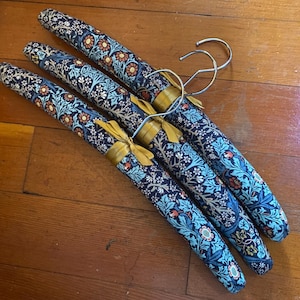 May include: Three padded clothes hangers covered in a dark blue fabric featuring a repeating floral pattern of orange, white, and light blue flowers with green leaves. Each hanger includes a silver metal hook and a small gold ribbon tied at the center.