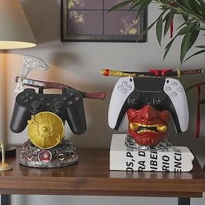 May include: Two gaming controller stands. The left stand features a silver axe and a gold circular shield. The right stand is a red and gold Japanese-style demon mask holding a white and black controller with a miniature katana resting on top.