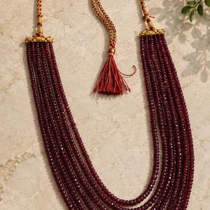 Op de afbeelding: Een meerstrengs ketting met vijf rijen kleine, gefacetteerde, donkerbordeauxrode kralen. De strengen zijn bevestigd met goudkleurige metalen kapjes en verbonden met een gevlochten goud en rood koord met een verstelbare schuifknoop en een rode kwast.