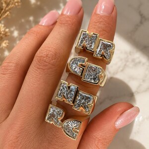 Puede incluir: Cuatro anillos dorados usados en los dedos, cada uno con letras grandes estilo bloque incrustadas con piedras claras facetadas. Los anillos muestran las letras NR, GB, NR y RC, con un acabado metálico pulido y un diseño grueso y rectangular.
