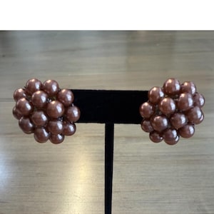 Op de afbeelding: Een paar cluster-oorbellen met meerdere ronde, metallic bruine kralen gerangschikt in een dichte, bolvormige formatie. Elke oorbel bestaat uit talloze kleine, glanzende bolletjes die samen een getextureerde, ronde vorm creëren.