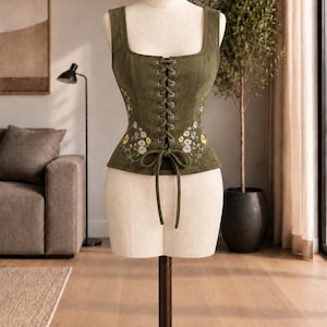 Può includere: Un corsetto color verde oliva con scollo quadrato, chiusura frontale con lacci in tessuto abbinato e delicati ricami floreali nei toni del giallo, bianco e blu sui pannelli laterali, esposto su un manichino da sartoria.