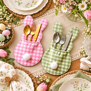 May include: Two fabric cutlery holders shaped like bunny heads with ears, one in pink and white gingham and one in green and white gingham. Each holder features a small white pom-pom tail and contains a gold or silver spoon, fork, and knife.