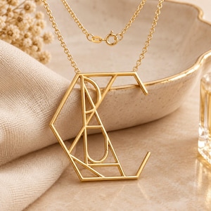 Op de afbeelding: Een goudkleurige ketting met een grote, geometrische hanger in de vorm van de letter C. De hanger is opgebouwd uit dunne, onderling verbonden metalen lijnen die een complexe, hoekige structuur vormen. Het hangt aan een fijne goudkleurige ketting.